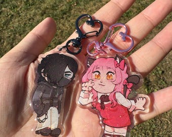 Puede incluir: Dos llaveros acrílicos con personajes de estilo anime. Uno muestra un personaje con una sudadera con capucha y máscara negras, el otro un personaje de pelo rosa con un vestido rojo. Ambos tienen cierres en forma de corazón.