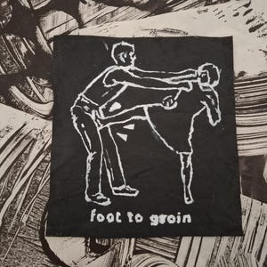 May include: Black square featuring a white illustration of two figures in a fighting pose. The words "foot to groin" are written in white at the bottom. The background has black and white abstract brushstrokes.
