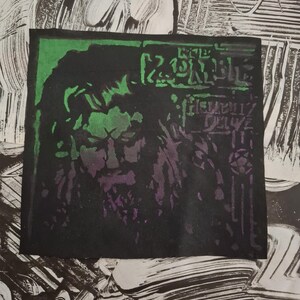 May include: A black and green patch featuring a stylized portrait of a person with long hair and a beard. The text "THE ZOMBIE" is visible at the top, with "DELUXE" below. The patch has a green border and purple accents.