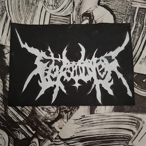 May include: A black rectangular patch with white, stylized text. The text appears to be a band name or logo, featuring sharp, angular lines and a gothic aesthetic. The background is a textured black and white pattern.