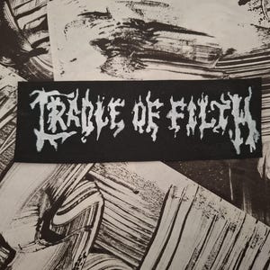 Op de afbeelding: Een zwart rechthoekig embleem met de witte tekst "CRADLE OF FILTH" in gotische letters. Het embleem is geplaatst tegen een achtergrond van abstracte zwart-witte penseelstreken.