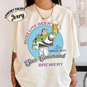 May include: Ivory-colored t-shirt featuring a graphic of Buzz Lightyear holding a beer, with the text "Buzz Lite Beer Star Command Brewery." The design includes a blue circle and the words "Best Quality Since 1995."