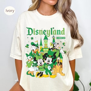 Puede incluir: Camiseta color marfil con un diseño de Disneyland para el Día de San Patricio. El diseño incluye a Mickey y Minnie Mouse, Donald y Daisy Duck, Goofy y Pluto, todos vestidos de verde. El logo de Disneyland es verde.