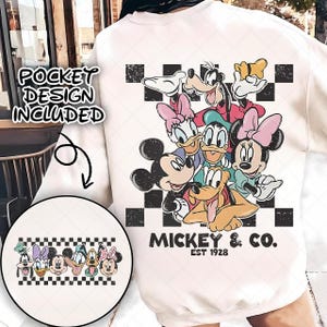 Puede incluir: Sudadera color crema con un gráfico de Mickey Mouse y sus amigos, incluyendo Minnie, Donald y Goofy, con un patrón de cuadros blancos y negros. El texto "Mickey & Co. EST 1928" está impreso debajo. Las palabras "Pocket Design Included" también son visibles.