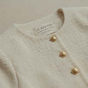 May include: Cream-colored tweed jacket with three gold-tone buttons. The jacket has a round neckline and a textured fabric. The label reads "LE NEUTRE MODERN CLASSIC & TIMELESS From here, with you."