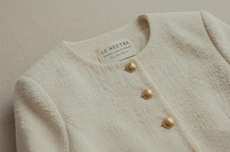 May include: Cream-colored tweed jacket with three gold-tone buttons. The jacket has a round neckline and a textured fabric. The label reads "LE NEUTRE MODERN CLASSIC & TIMELESS From here, with you."