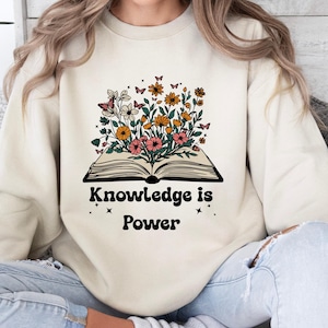 May include: Cream-coloured sweatshirt featuring a graphic of an open book with flowers and butterflies. The text "Knowledge is Power" is printed below. The sweatshirt has a crew neck and is a neutral colour.