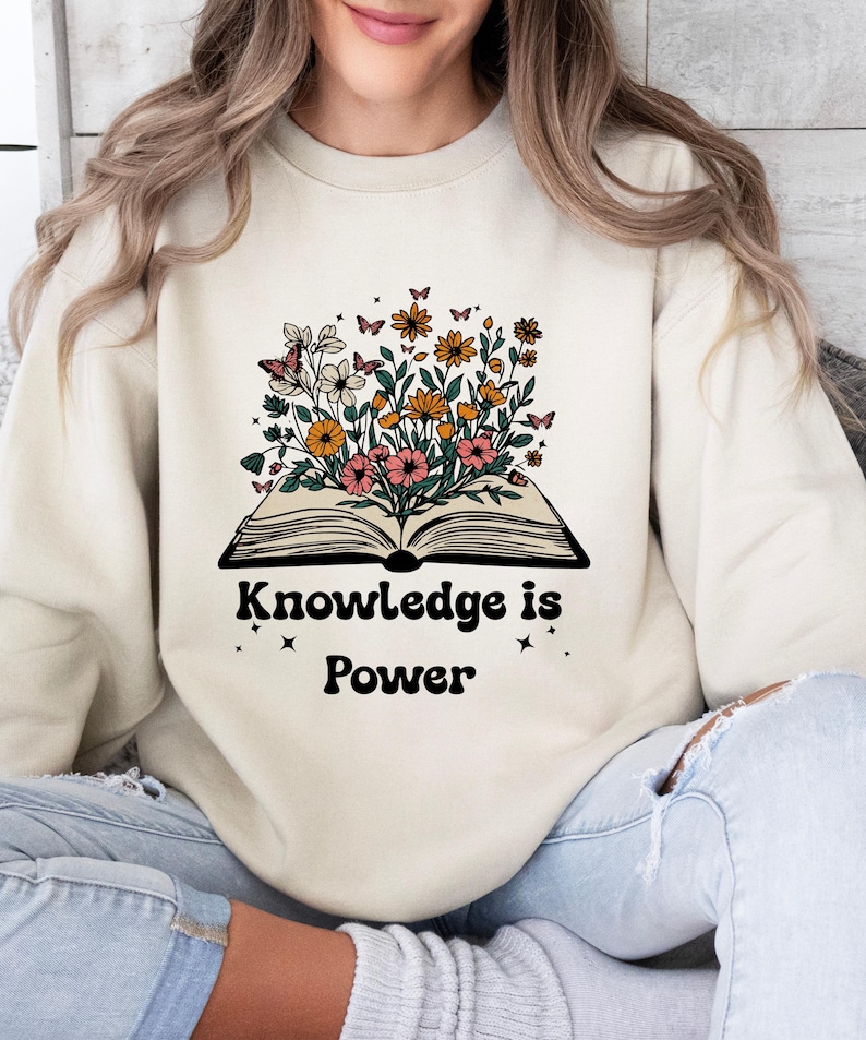 May include: Cream-coloured sweatshirt featuring a graphic of an open book with flowers and butterflies. The text "Knowledge is Power" is printed below. The sweatshirt has a crew neck and is a neutral colour.