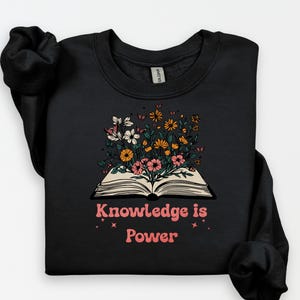 May include: Black sweatshirt with an open book design, from which colourful flowers are blossoming. The pink text "Knowledge is Power" is below the book, with star accents. The sweatshirt has a crew neck and long sleeves.