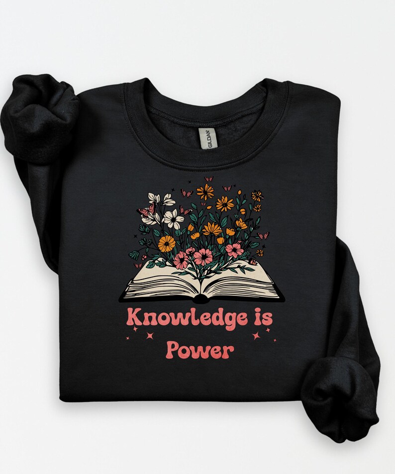 May include: Black sweatshirt with an open book design, from which colourful flowers are blossoming. The pink text "Knowledge is Power" is below the book, with star accents. The sweatshirt has a crew neck and long sleeves.