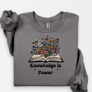 May include: A grey sweatshirt with an open book design, from which colourful flowers and butterflies emerge. The text "Knowledge is Power" is printed below the book. The sweatshirt has long sleeves.