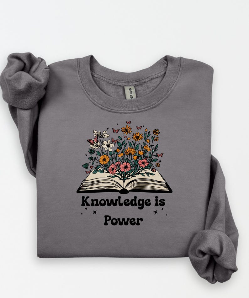 May include: A grey sweatshirt with an open book design, from which colourful flowers and butterflies emerge. The text "Knowledge is Power" is printed below the book. The sweatshirt has long sleeves.