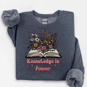 May include: Dark grey sweatshirt with the text "Knowledge is Power" in pink. The sweatshirt features an open book graphic with colourful flowers and butterflies. The Gildan brand tag is visible.