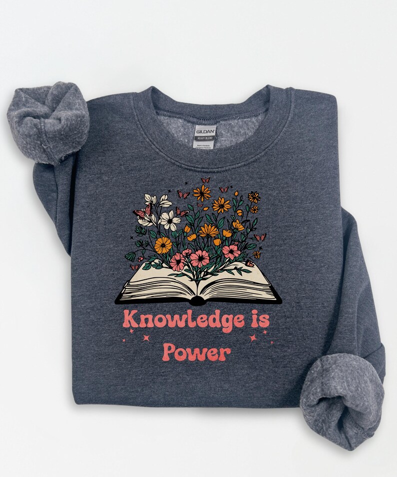 May include: Dark grey sweatshirt with the text "Knowledge is Power" in pink. The sweatshirt features an open book graphic with colourful flowers and butterflies. The Gildan brand tag is visible.