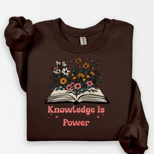 May include: A brown sweatshirt with a floral design sprouting from an open book. The design features white, pink, and yellow flowers, with the text "Knowledge is Power" in pink. The sweatshirt has a crew neck.