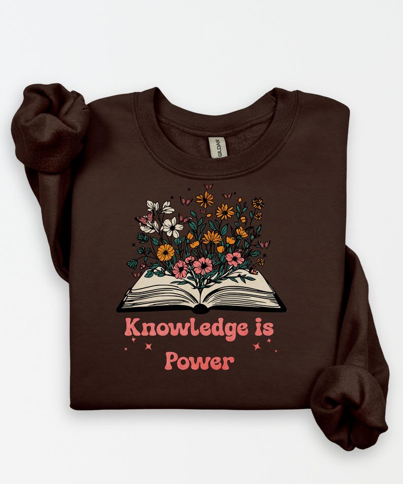 May include: A brown sweatshirt with a floral design sprouting from an open book. The design features white, pink, and yellow flowers, with the text "Knowledge is Power" in pink. The sweatshirt has a crew neck.