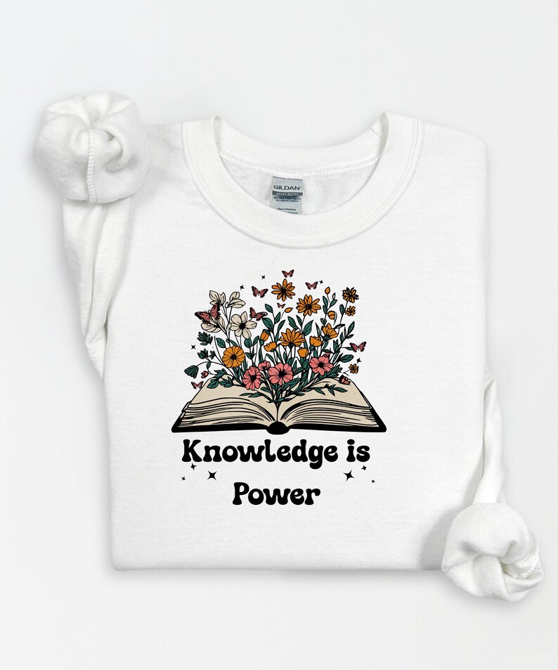 May include: White sweatshirt with a floral design. The graphic features an open book with colourful flowers and butterflies emerging from the pages. The text "Knowledge is Power" is below the book.