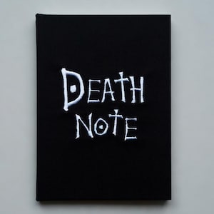 May include: Black notebook with the words "Death Note" in white, stylized lettering. The notebook is rectangular and appears to be closed, with a plain, solid black cover. The background is a light gray.