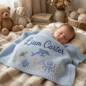 Peut inclure: Une couverture pour bébé en tricot bleu clair avec le nom Liam Carter brodé en écriture cursive bleu foncé. La couverture présente des motifs de vie marine incluant un dauphin, une tortue, un hippocampe, une pieuvre, une étoile de mer et des coquillages.