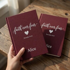 以下が含まれることがあります： 「faith over fear」の筆記体、ハート、そして「Proverbs 18:24」が表紙に印刷された、2冊のバーガンディ色のジャーナル。1冊は「Alice」という名前でパーソナライズされています。ジャーナルは木製の表面に置かれています。