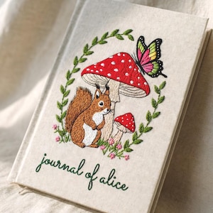 Pode incluir: Um diário bege com um desenho bordado com um esquilo, cogumelos e uma borboleta. A capa tem as palavras "journal of alice" em verde. O desenho é rodeado por uma borda de folhas verdes.