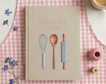 May include: A beige cookbook titled "Anabelle's Favourite Recipes" with embroidered kitchen tools: a whisk, wooden spoon, and rolling pin. The book rests on a pink gingham apron, with a cup of coffee and flowers nearby.