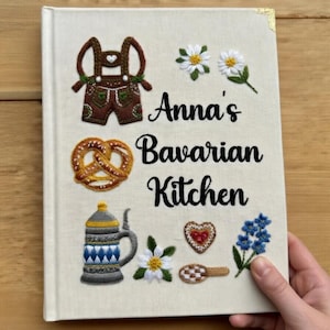Könnte beinhalten: Ein leinenbezogenes Kochbuch mit dem Titel "Anna's Bavarian Kitchen" mit gestickten bayerischen Motiven. Das Cover zeigt eine Brezel, einen Bierkrug und florale Akzente. Das Buch hat goldene Eckenschützer.