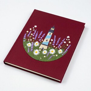 May include: A burgundy journal featuring an embroidered design of a lighthouse, daisies, and lavender. The lighthouse is white and blue, with a black top. The flowers are white and purple, set against a green background.