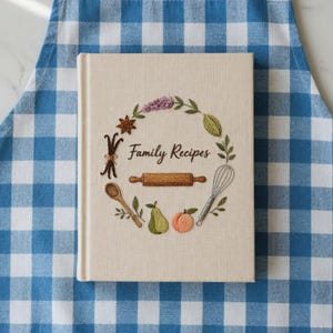 May include: A beige recipe book with the words "Family Recipes" in script. The cover features an embroidered wreath with a rolling pin, whisk, pear, peach, and other kitchen-themed elements. The book is on a blue and white checkered apron.
