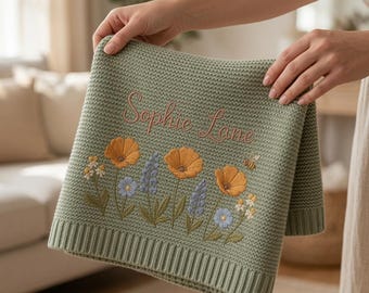 May include: A sage green knit baby blanket featuring the embroidered name Sophie Lane in a cursive font. The blanket is decorated with a floral pattern of orange poppies, blue lupines, and small white flowers, along with a small embroidered bee.