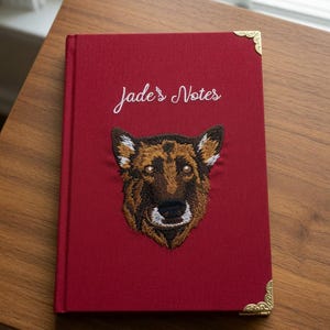 May include: A deep red hardback journal featuring an embroidered German Shepherd dog face on the front cover. The text "Jade's Notes" is stitched in white above the dog. The journal includes decorative gold-toned metal corner protectors.