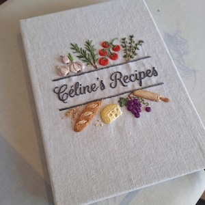 Może przedstawiać: Lniana książka kucharska z haftowanymi detalami. Okładka zawiera napis "Céline's Recipes" nad haftowanym czosnkiem, pomidorami, ziołami, bagietką, serem, winogronami i wałkiem do ciasta. Książka jest prostokątna i wydaje się zamknięta.
