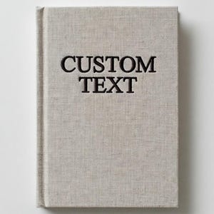 Può includere: Un libro con copertina rigida con una copertina in lino grigio chiaro. Le parole "CUSTOM TEXT" sono ricamate in nero sul davanti. Il libro è rettangolare e sembra vuoto all'interno, pronto per contenuti personalizzati.
