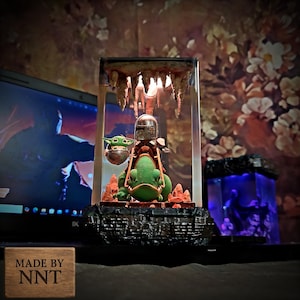 Puede incluir: Una l&aacute;mpara decorativa con una figura mandaloriana montada en una criatura verde, con una figura de Baby Yoda en una c&aacute;psula plateada. La l&aacute;mpara est&aacute; encerrada en una caja transparente con una base rocosa y una l&aacute;mpara. La base tiene el texto "MADE BY NNT".