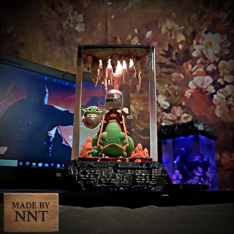 Puede incluir: Una l&aacute;mpara decorativa con una figura mandaloriana montada en una criatura verde, con una figura de Baby Yoda en una c&aacute;psula plateada. La l&aacute;mpara est&aacute; encerrada en una caja transparente con una base rocosa y una l&aacute;mpara. La base tiene el texto "MADE BY NNT".