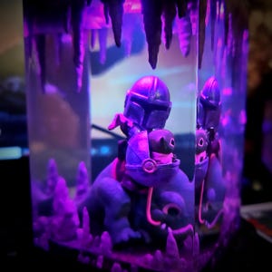 Puede incluir: Una figura de Mandaloriano montando una criatura azul, encerrada en una vitrina rectangular transparente. La escena est&aacute; iluminada con luz p&uacute;rpura, creando un efecto dram&aacute;tico. La vitrina tiene una estructura similar a una cueva en la parte superior.