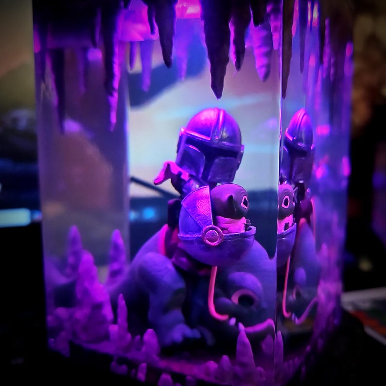 Puede incluir: Una figura de Mandaloriano montando una criatura azul, encerrada en una vitrina rectangular transparente. La escena est&aacute; iluminada con luz p&uacute;rpura, creando un efecto dram&aacute;tico. La vitrina tiene una estructura similar a una cueva en la parte superior.
