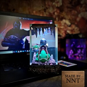 Puede incluir: Escultura de resina iluminada con The Mandalorian y Grogu montados en una criatura parecida a una rana. La escultura est&aacute; dentro de un bloque transparente con una parte superior en forma de cueva e iluminaci&oacute;n azul. Una placa de madera con "MADE BY NNT" es visible.
