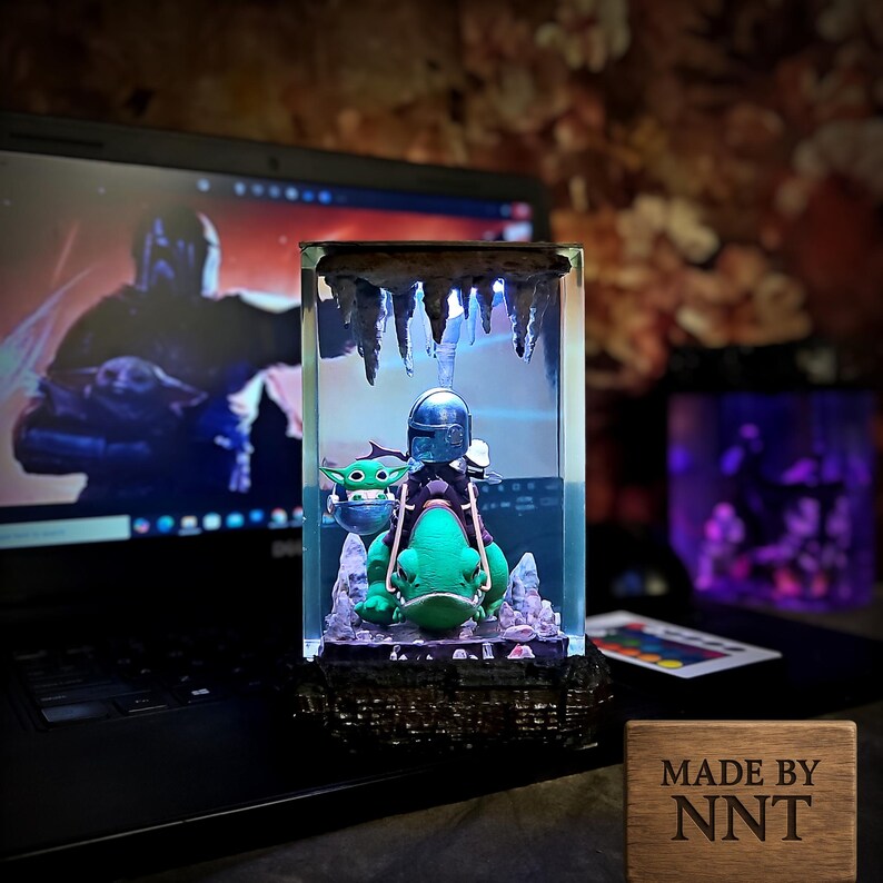 Puede incluir: Escultura de resina iluminada con The Mandalorian y Grogu montados en una criatura parecida a una rana. La escultura est&aacute; dentro de un bloque transparente con una parte superior en forma de cueva e iluminaci&oacute;n azul. Una placa de madera con "MADE BY NNT" es visible.