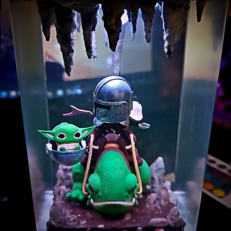 Puede incluir: Figura detallada de un personaje mandaloriano montado en una criatura verde, con un peque&ntilde;o alien&iacute;gena verde en un portabeb&eacute;s flotante. La escena est&aacute; ambientada en un recipiente transparente con una estructura similar a una cueva en la parte superior.