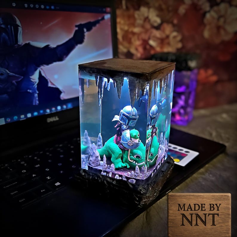 Puede incluir: Cubo decorativo de resina con una escena de figuras montadas en criaturas verdes, iluminado con luz azul. El cubo tiene una parte superior de madera y una base texturizada. El texto "MADE BY NNT" est&aacute; en una placa de madera.