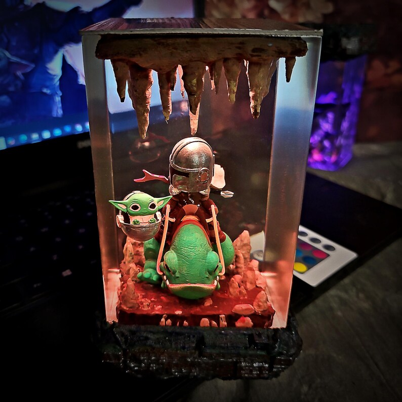 Puede incluir: Diorama decorativo de resina que presenta una figura mandaloriana montada en una criatura verde con forma de rana, con Grogu en un portabeb&eacute;s. La escena est&aacute; ambientada en un entorno similar a una cueva con estalactitas e iluminaci&oacute;n roja.