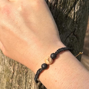 May include: Black braided bracelet with three beads. Two are dark, textured, and spherical, and the center bead is light-colored with a carved symbol. The bracelet is worn on a wrist.