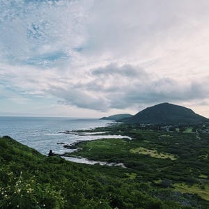 May include: A scenic coastal landscape featuring a vast expanse of ocean meeting a lush, green shoreline. The sky is filled with a mix of white and gray clouds. A person is sitting on the hillside, enjoying the view.