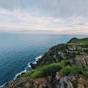 May include: A scenic coastal landscape featuring a vast blue ocean under a cloudy sky. The foreground shows a rocky cliff covered in green grass, with the ocean waves gently crashing against the shore.