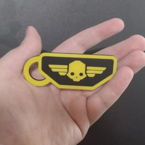 May include: A yellow and black patch in the shape of a coffee cup with a skull and wings design. The patch is held in a hand against a dark background.