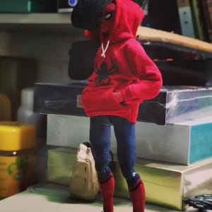 May include: A Spider-Man action figure wearing a red hoodie with a black spider emblem and white drawstrings. The figure has blue jeans and red and black boots. The figure is standing with its hands in the hoodie pockets.