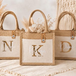 Könnte beinhalten: Drei Jute-Tragetaschen mit personalisierten Namen: "Nicole", "Kinga" und "Diane". Jede Tasche hat eine natürliche Jute-Außenseite, eine cremefarbene Vordertasche und einen passenden Griff. Trockenblumen und Etiketten mit QR-Codes sind sichtbar.