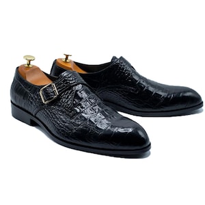 May include: A pair of black leather dress shoes with a crocodile texture. The shoes have a single strap with a gold-coloured buckle. Wooden shoe trees are inside the shoes.