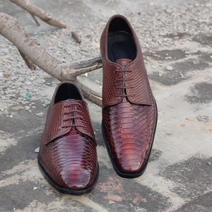 Peut inclure: Une paire de chaussures de ville en cuir marron avec un motif texturé en forme de peau de serpent. Les chaussures ont un design classique à lacets et une finition polie. Les chaussures sont présentées sur une surface en briques.